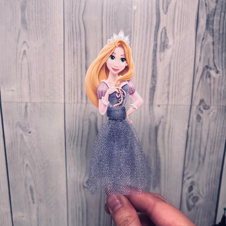 May include: A princess-themed cake topper featuring a cartoon illustration of a princess with long blonde hair, a silver crown, and a purple and gray dress. The princess is holding her hand on her hip. The dress has a tulle skirt.