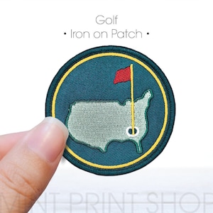 以下が含まれることがあります： ゴルフをテーマにした丸いアイロン接着パッチ。パッチには、赤い旗が黄色いポールに立っている、緑色の米国地図が描かれています。「Golf - Iron on Patch」というテキストがパッチの上にあります。