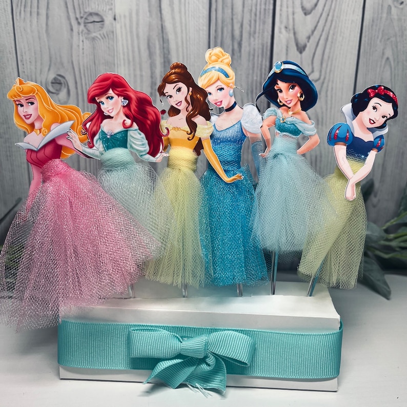 May include: A collection of six princess-themed cake toppers. Each princess has a unique dress color, including pink, teal, yellow, blue, and white. The toppers are attached to a white base with a light blue ribbon and bow.
