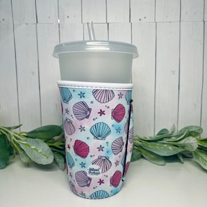 May include: A reusable tumbler with a lid and straw, encased in a patterned sleeve. The sleeve features a design of pink, blue, and white seashells and starfish on a watercolor background. The text "Mind Print" is visible on the sleeve.