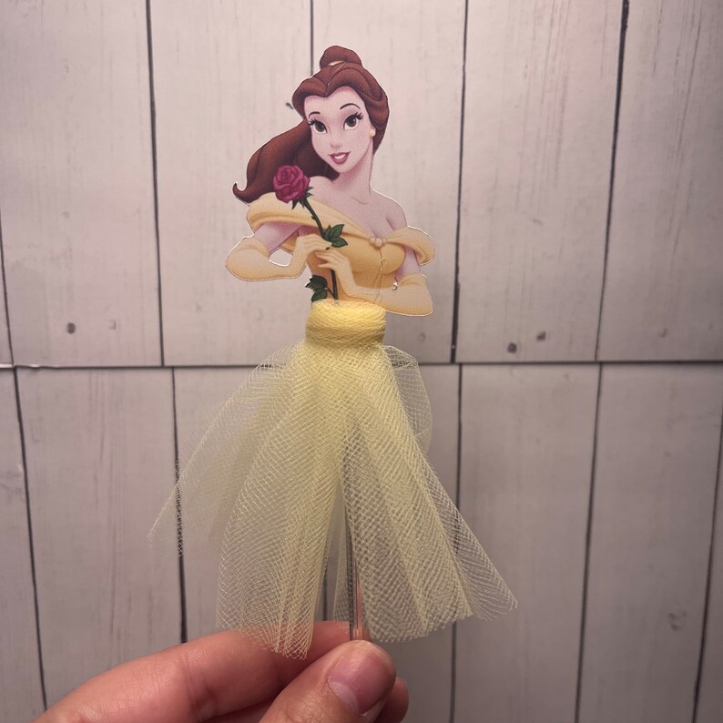 May include: A Belle-themed cake topper featuring a cartoon illustration of Belle in a yellow gown, holding a red rose. The dress has a tulle skirt. The topper is attached to a silver wire.