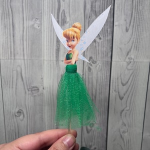 May include: A Tinkerbell-themed cake topper featuring a cartoon image of the fairy with a green tulle skirt and white wings. The fairy has blonde hair and is wearing a green top. The topper is attached to a wooden stick.