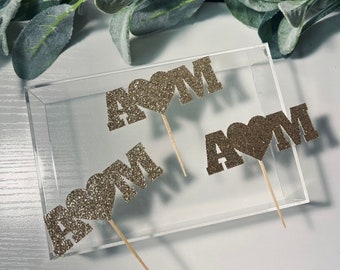 Pode incluir: Três toppers de bolo de glitter dourado com a palavra "MOM" e um coração no centro de cada topper. Os toppers estão em uma superfície de acrílico transparente.