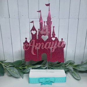 May include: Pink glitter castle cake topper with the name "Amaya" on the front. The castle is on a white base with a turquoise ribbon and bow.