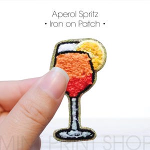 May include: Iron-on patch of an Aperol Spritz cocktail in a glass with an orange slice. The drink is orange, red, and yellow with a white rim. The glass is outlined in gold glitter.