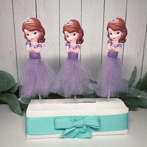 Peut inclure: Trois décorations de gâteau sur le thème des princesses. Chaque figurine représente une princesse de dessin animé avec une tiare, les bras croisés et une jupe en tulle violet. Elles sont fixées sur une boîte blanche avec un ruban et un nœud turquoise.