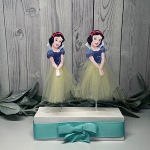 May include: Two Snow White character toppers with yellow tulle skirts and blue and white tops are displayed on a white base with a teal ribbon and bow. The background is a light gray wooden surface.