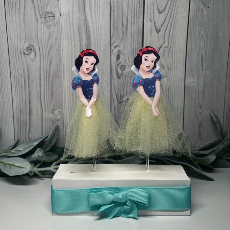 May include: Two Snow White character toppers with yellow tulle skirts and blue and white tops are displayed on a white base with a teal ribbon and bow. The background is a light gray wooden surface.
