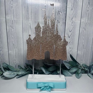 May include: Glittery gold castle cake topper with two flags atop the towers. The castle is mounted on a white base with a light blue ribbon and bow. The background is a light wood grain.