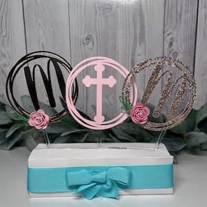 May include: Three round cake toppers with a cross, the letter "M" and a floral design. The toppers are on a white box with a blue ribbon.