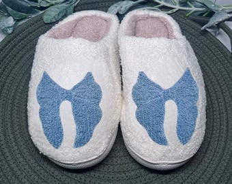 May include: A pair of white, fluffy slippers with a light blue bow design on the front. The interior is a soft pink color. The slippers are designed for indoor use and are placed on a green surface.