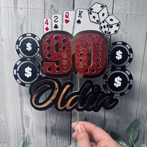 May include: A red and gold glitter cake topper with the number 90 and the word "Oldin" in black and gold glitter. The topper also features playing cards, dice, and poker chips with dollar signs.