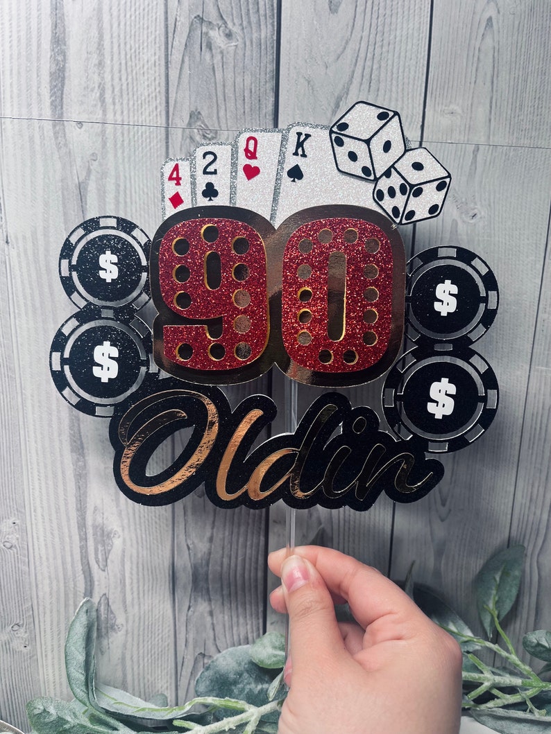 May include: A red and gold glitter cake topper with the number 90 and the word "Oldin" in black and gold glitter. The topper also features playing cards, dice, and poker chips with dollar signs.