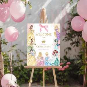 May include: A wooden easel displays a pink and gold welcome sign for a princess-themed birthday party. The sign features illustrations of princesses and the text "Welcome to Princess Charlotte's Fifth Birthday."