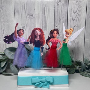 May include: A decorative centerpiece featuring four cartoon character figures with tulle skirts in purple, blue, red, and green. The figures are mounted on a white base with a teal ribbon and bow.