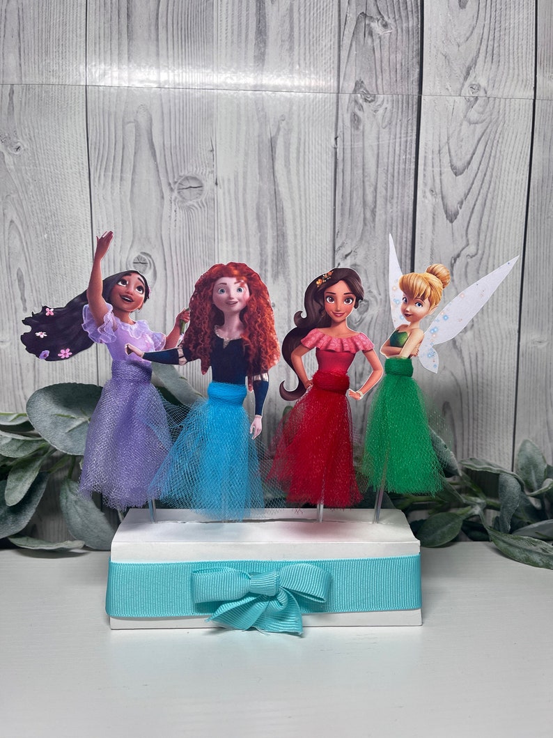 May include: A decorative centerpiece featuring four cartoon character figures with tulle skirts in purple, blue, red, and green. The figures are mounted on a white base with a teal ribbon and bow.