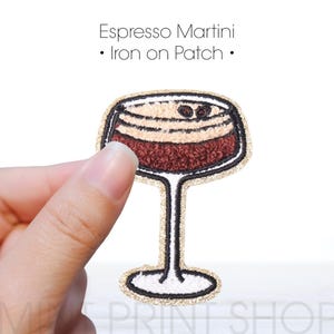Könnte beinhalten: Espresso Martini Bügeleisen-Aufnäher mit goldener Umrandung und weißem Hintergrund. Der Aufnäher zeigt ein braunes und beiges Martini-Glas mit Kaffebohnen darauf.