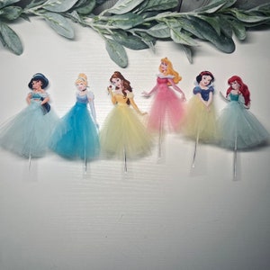 May include: Six princess-themed cupcake toppers. Each princess is depicted in a colorful gown with a tulle skirt, mounted on a clear stick. The princesses include Jasmine, Cinderella, Belle, Aurora, Snow White, and Ariel.