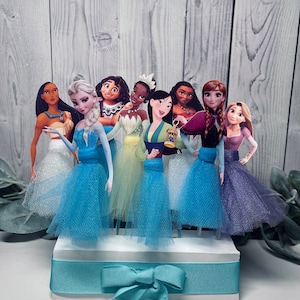 May include: A collection of princess-themed cake toppers. Each princess is depicted in a colorful gown with a tulle skirt. The toppers are arranged on a white base with a light blue ribbon and bow. The background is a light wood-grain pattern.