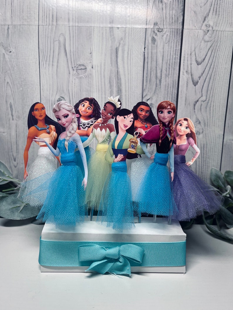 May include: A collection of princess-themed cake toppers. Each princess is depicted in a colorful gown with a tulle skirt. The toppers are arranged on a white base with a light blue ribbon and bow. The background is a light wood-grain pattern.