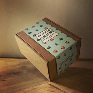 Peut inclure: Une boîte en carton marron avec une étiquette à motifs bleus et verts. L'étiquette indique "Patakid" et "Fabrique toi-même ta pâte à modeler !"