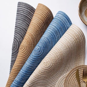 May include: A collection of woven placemats in various colors, including gray, tan, blue, and beige. The placemats have a circular design with concentric rings, and a matching plate with a fork and knife is visible.