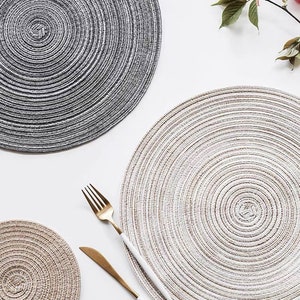 May include: A top-down view showcases a collection of woven placemats in varying sizes and colors. The largest placemat is a light beige, while another is a darker gray. A gold fork and knife with white handles are placed on the beige placemat. The theme is dining and home decor.