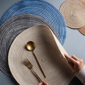 May include: A collection of round placemats in various colors, including blue, gray, and beige. The placemats have a woven texture and are arranged on a gray surface. A gold spoon and fork are placed on one of the beige placemats.