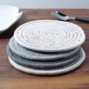 May include: A set of three round woven coasters. The top coaster is white with a colorful rainbow pattern. The bottom two coasters are gray.