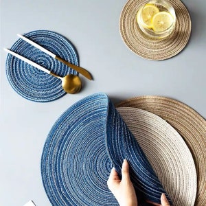 May include: Circular placemats in shades of blue, beige, and brown, with a textured, woven design. A set of gold and white utensils rests on a blue placemat. A glass of water with lemon slices sits on a beige placemat. The placemats are arranged on a gray surface.