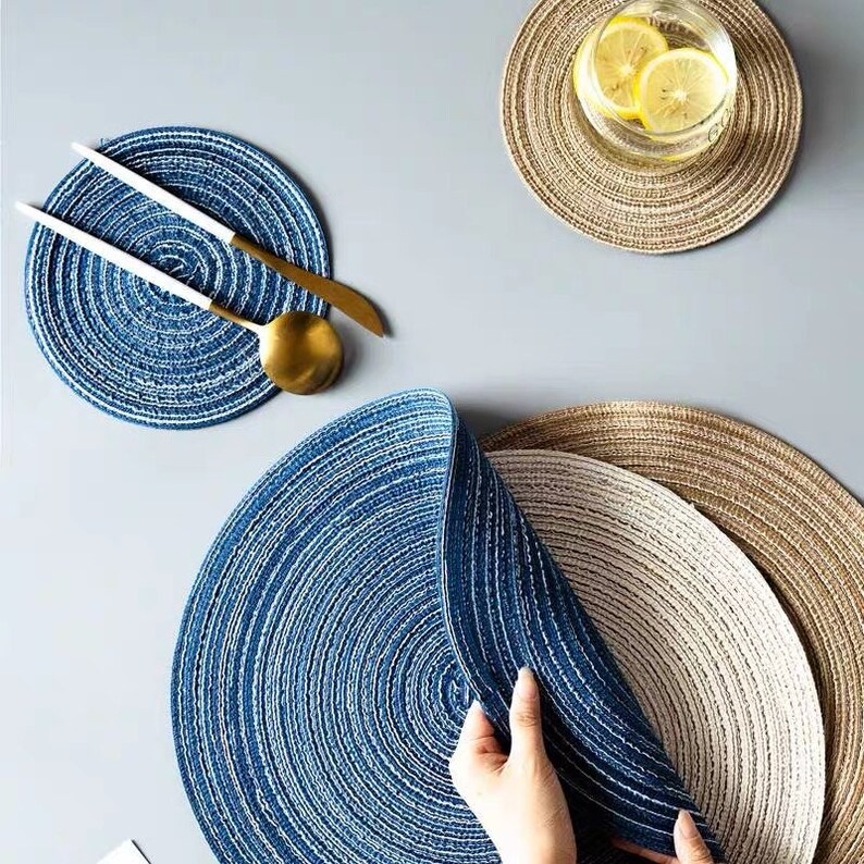 May include: Circular placemats in shades of blue, beige, and brown, with a textured, woven design. A set of gold and white utensils rests on a blue placemat. A glass of water with lemon slices sits on a beige placemat. The placemats are arranged on a gray surface.