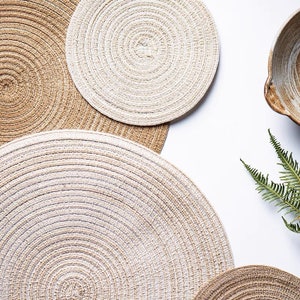 May include: Overhead shot of several round woven placemats in shades of beige and brown. The placemats have a concentric circle design. A ceramic bowl and a sprig of green fern are also visible.