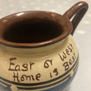 May include: A brown and white ceramic mug with a blue and brown design. The mug has a handle and says "East or West Home is Best" on the side.