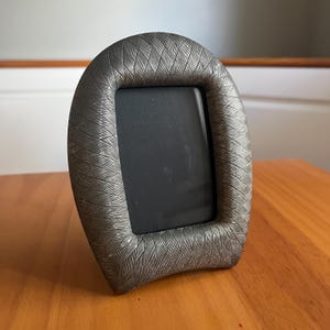 May include: A freestanding picture frame with a rounded top and a rectangular center opening. The frame features a textured, metallic gray finish with a repeating diagonal cross-hatch pattern that covers the entire surface of the border.