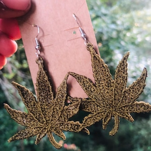 May include: A pair of gold-colored leaf-shaped earrings with silver hooks. The earrings are held against a brown tag, with a blurred green background. The leaves have detailed stitching.