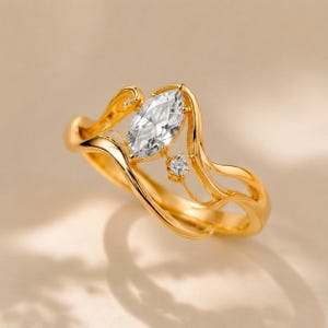 May include: A gold ring featuring a marquise-cut clear gemstone as the centrepiece, accented by a single small round clear gemstone. The band has a fluid, wavy design that curves around the stones, creating an open, asymmetrical silhouette.