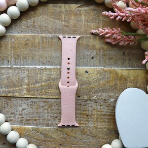 May include: A pink silicone Apple Watch band with a repeating pattern of small hearts. The band is on a wooden background with a white heart and a string of wooden beads.