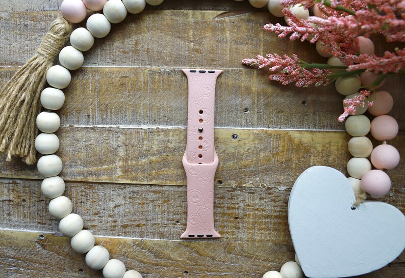 May include: A pink silicone Apple Watch band with a repeating pattern of small hearts. The band is on a wooden background with a white heart and a string of wooden beads.