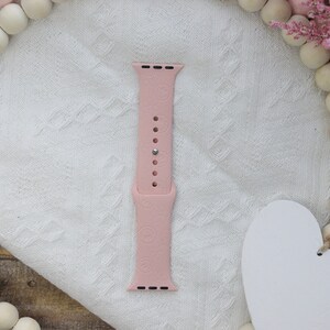 May include: A pink silicone Apple Watch band with a floral pattern. The band is shown on a white fabric background.