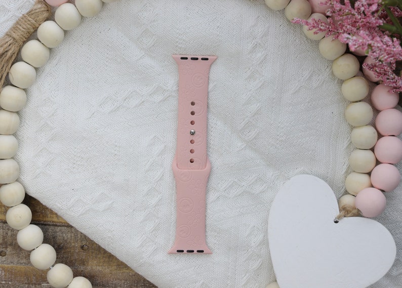 May include: A pink silicone Apple Watch band with a floral pattern. The band is shown on a white fabric background.