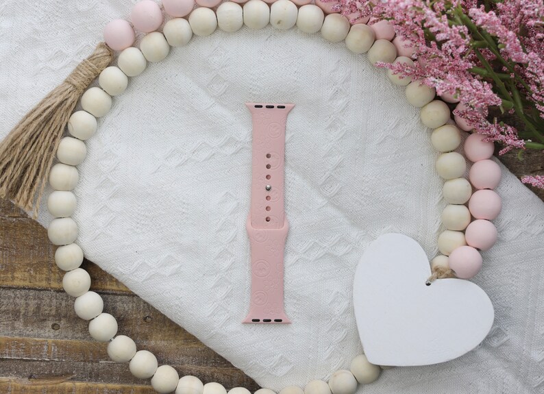 May include: A pink silicone watch band with a floral pattern. The band is on a white surface with a wooden bead garland.