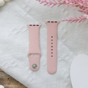 May include: Two pink silicone Apple Watch bands with a repeating pattern of small circles. The bands are laid out flat on a white fabric surface.