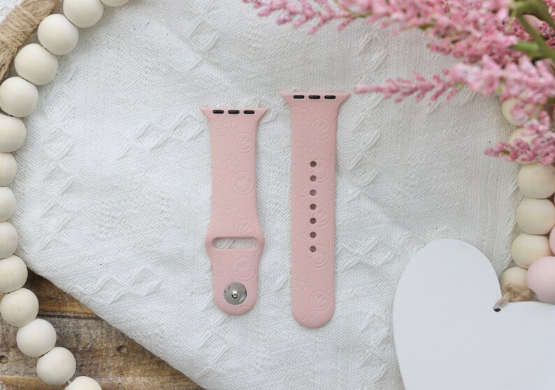 May include: Two pink silicone Apple Watch bands with a repeating pattern of small circles. The bands are laid out flat on a white fabric surface.