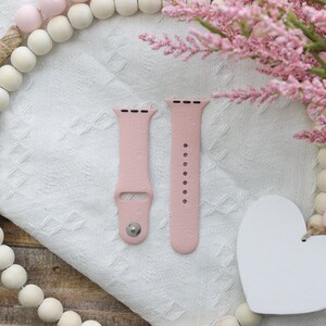 May include: Two pink silicone watch bands with a textured pattern. The bands are on a white surface with a pink and white beaded garland and a white heart.