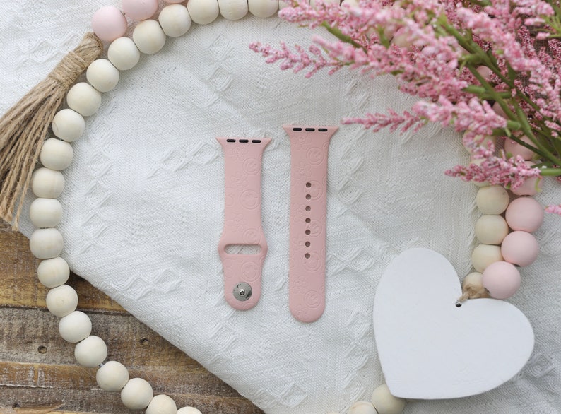 May include: Two pink silicone watch bands with a textured pattern. The bands are on a white surface with a pink and white beaded garland and a white heart.