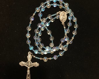 May include: A silver rosary with iridescent blue beads and a silver crucifix. The centerpiece features a silver oval with an image of Jesus.