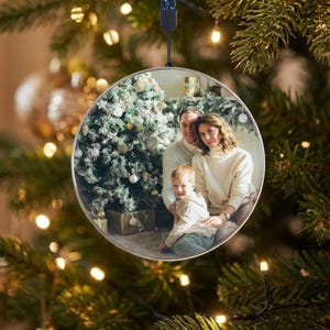 May include: A clear, round Christmas ornament with a family photo inside, hanging from a black cord. The photo shows a family of three in front of a decorated Christmas tree. The ornament is surrounded by a blurred background of Christmas lights and greenery.