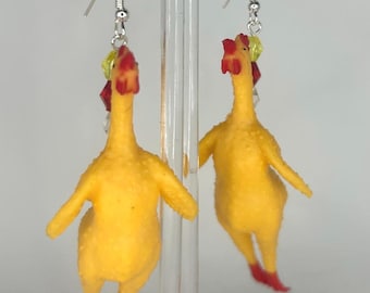 May include: A pair of dangle earrings featuring yellow rubber chickens with red feet and beaks. The chickens are hanging from silver hooks with green and red beads.