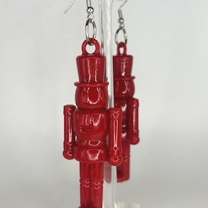 May include: A pair of red nutcracker earrings with silver hooks. The nutcrackers are wearing tall hats and have a detailed design.