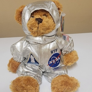 Puede incluir: Un oso de peluche marrón que lleva un traje de astronauta plateado con parches de la NASA y RTF. El oso está sentado sobre una superficie blanca.
